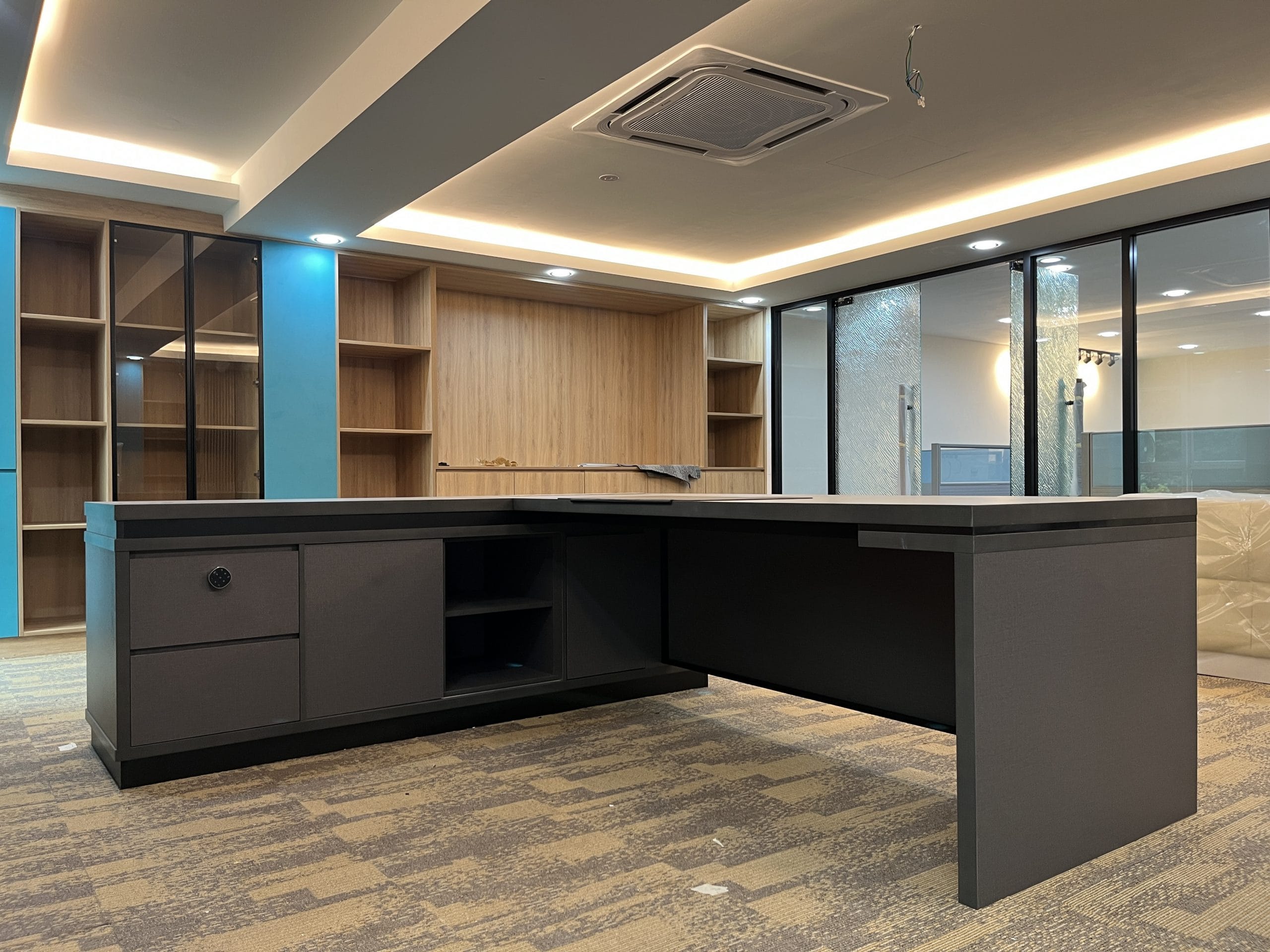 An open office area with built-in open shelving and a timber pantry run along the back wall, and a loose dark L-shaped desk with a side cabinet in the foreground.
