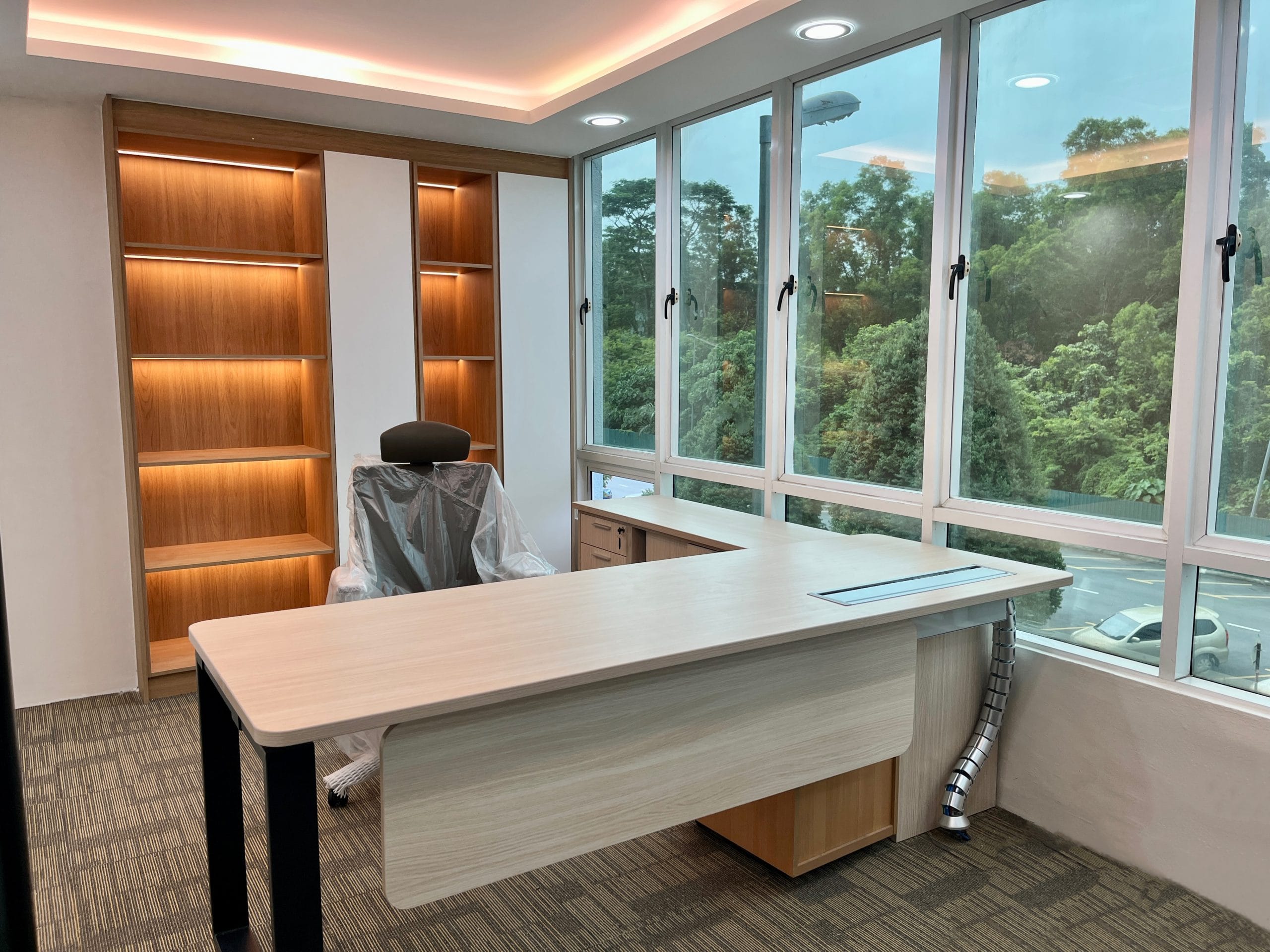 A director's room near handover: a built-in lit timber bookcase against the back wall, a freestanding curved-top L-desk in front of it, and the task chair still wrapped in protective plastic.