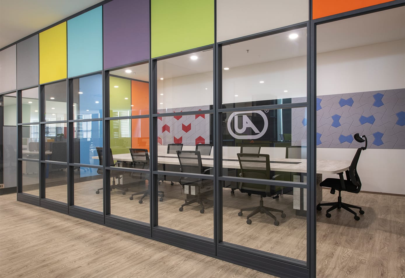 Glass-partitioned meeting room in the showroom carrying the UA logo, with a meeting table, mesh chairs and coloured glazing panels in the framing.