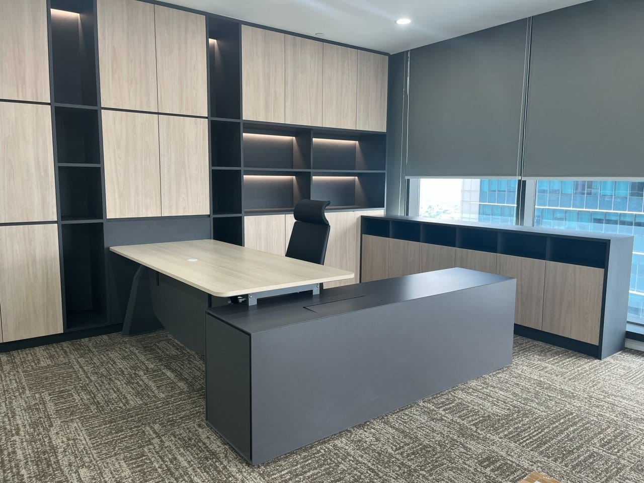 An executive room with a full-height built-in cabinet wall in pale timber and dark grey, including lit open display niches, with a loose L-shaped desk and a matching low credenza in front of it.